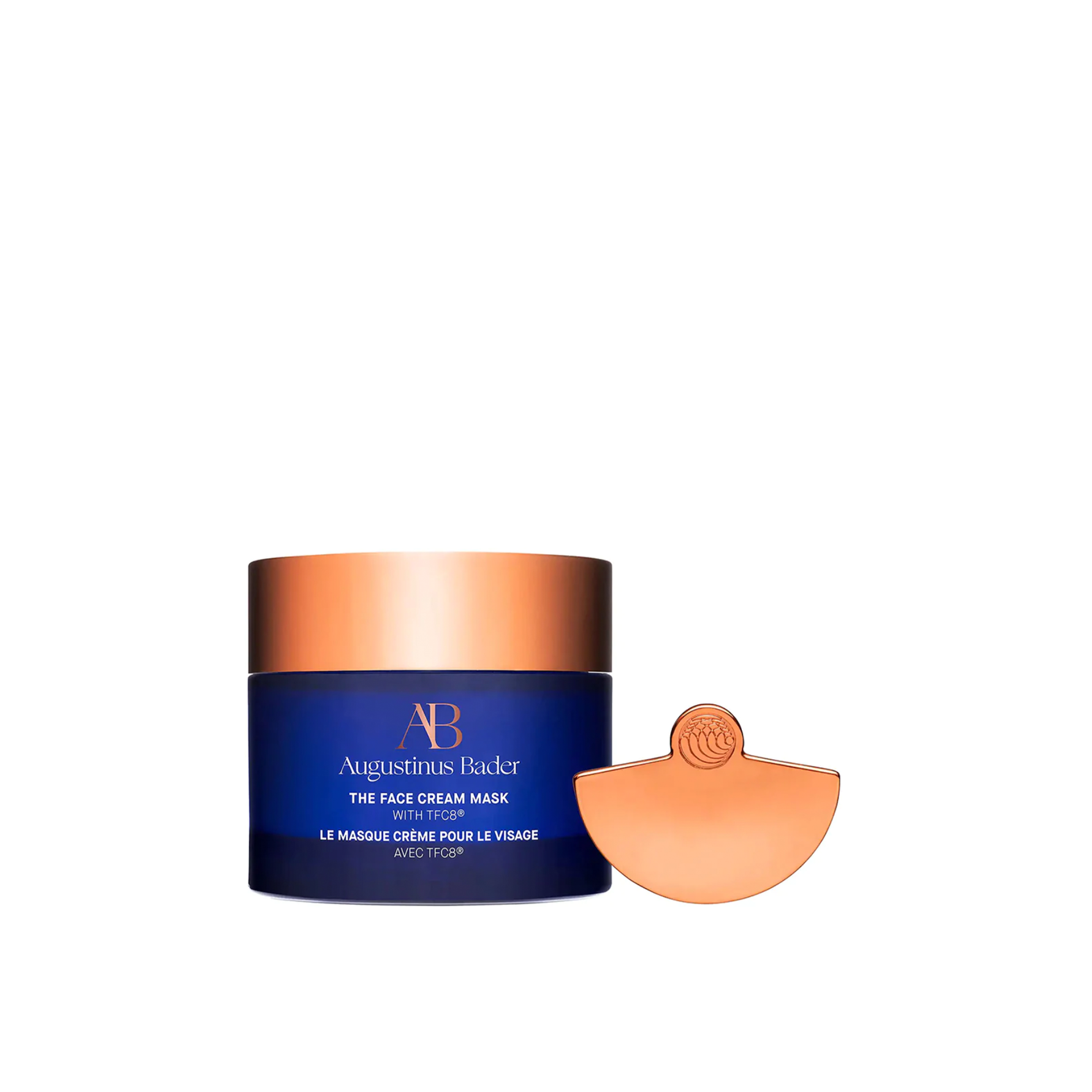 A jar of Augustinus Bader The Face Cream Mask with a copper lid and label, next to a copper-colored applicator.