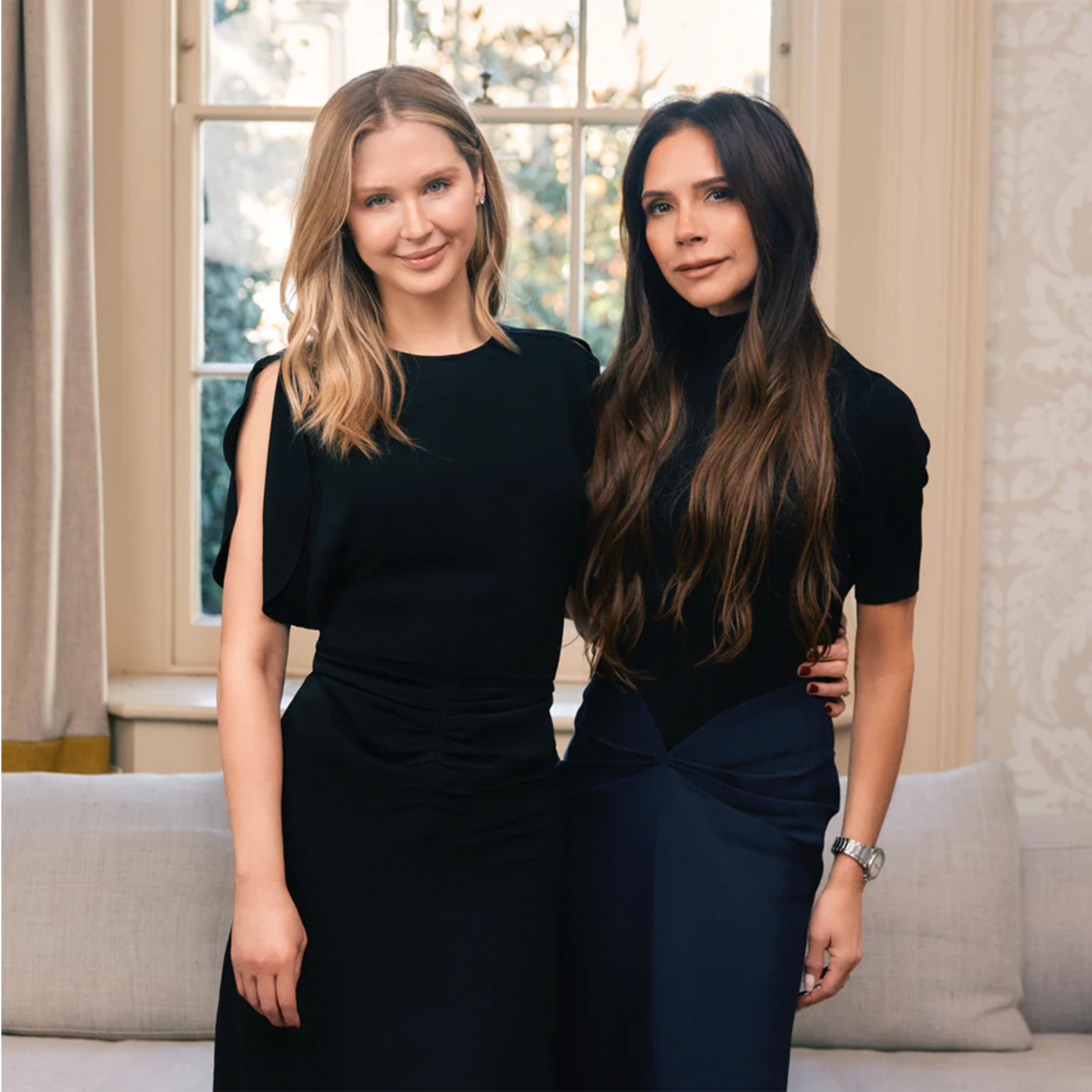 Two women in black dresses standing close together indoors with a window in the background.