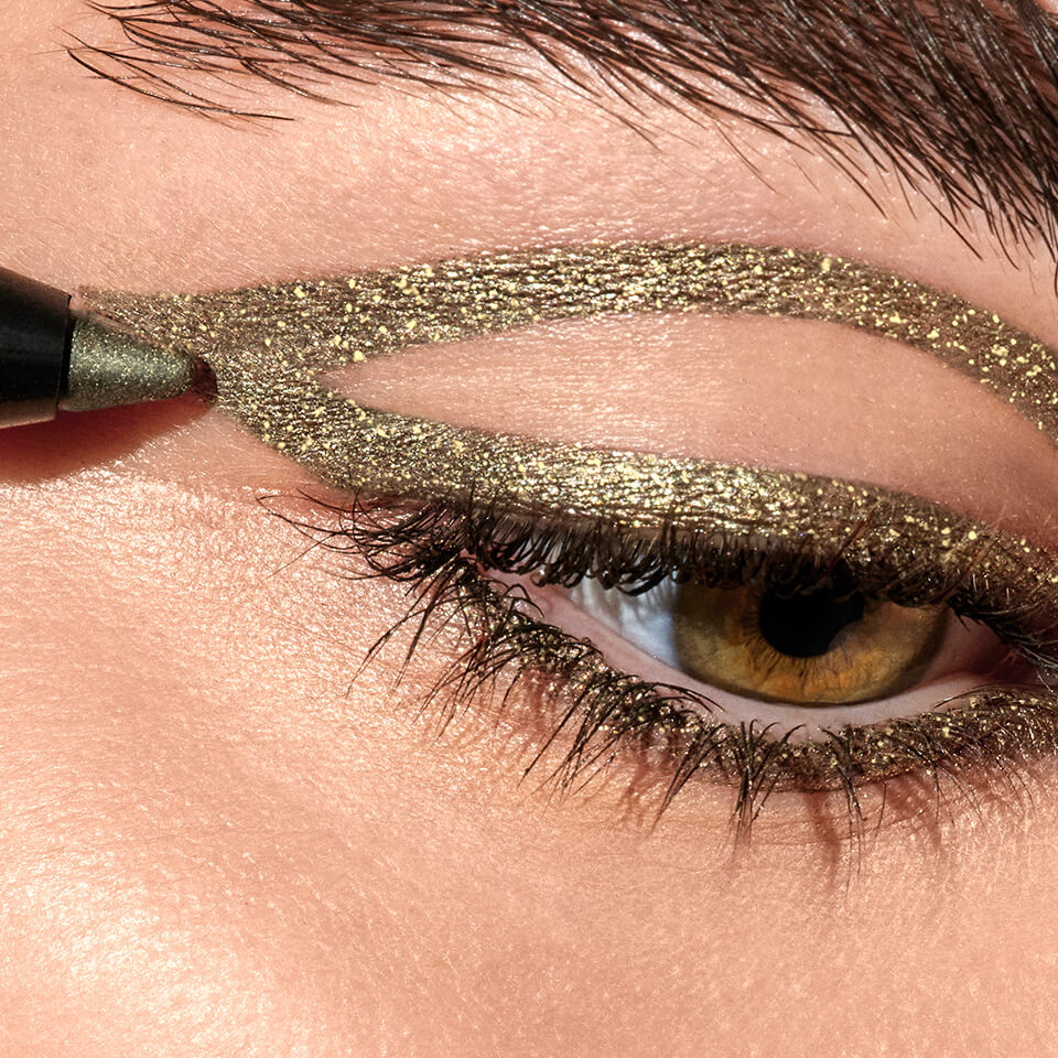 A close up image of an eye with gold sparkly eyeliner being applied to the eyelid.