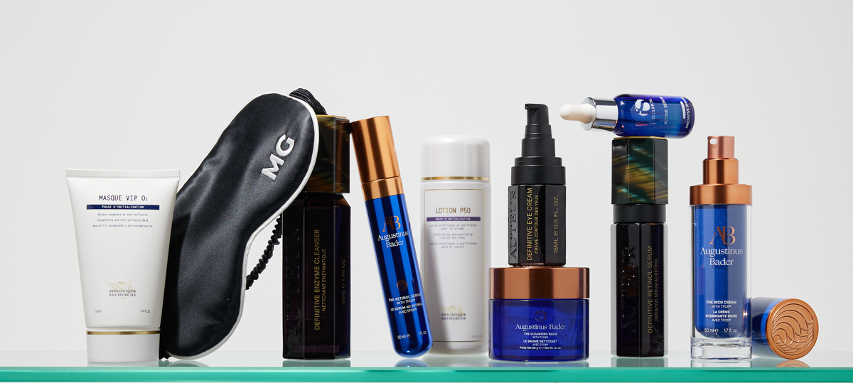 Skincare bottles, a silk MG sleep mask and a red travel case on a glass shelf