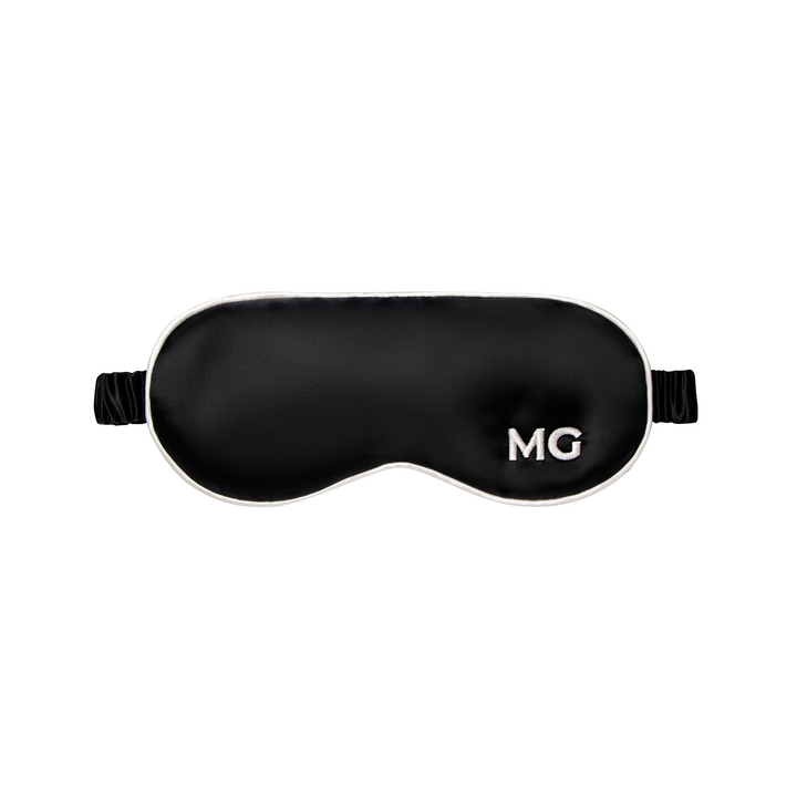 A black silk sleep mask with the initials 'MG' embroidered on the front.