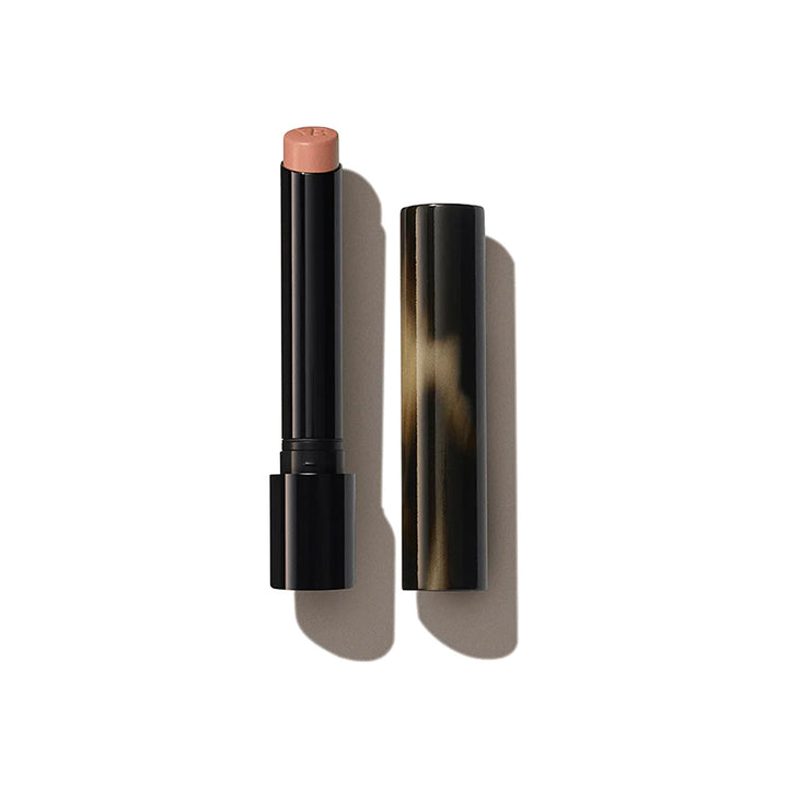 A Victoria Beckham Beauty Posh Lipstick in a light neutral pink shade, with the cap removed, displayed against a shadowed background.